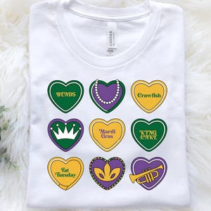 Peut inclure: T-shirt blanc avec neuf motifs en forme de c&oelig;ur repr&eacute;sentant des mots et des images sur le th&egrave;me du Mardi Gras. Les mots incluent "Beads", "Crawfish", "King Cake", "Mardi Gras", "Fat Tuesday", et "Trumpet".