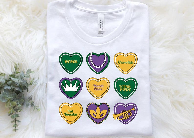 Peut inclure: T-shirt blanc avec neuf motifs en forme de c&oelig;ur repr&eacute;sentant des mots et des images sur le th&egrave;me du Mardi Gras. Les mots incluent "Beads", "Crawfish", "King Cake", "Mardi Gras", "Fat Tuesday", et "Trumpet".