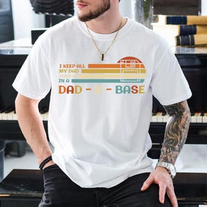 Puede incluir: Camiseta blanca con un gr&aacute;fico de estilo retro que dice "I keep all my dad jokes in a dad-a-base". El gr&aacute;fico presenta una cinta de casete y un esquema de color arco&iacute;ris.