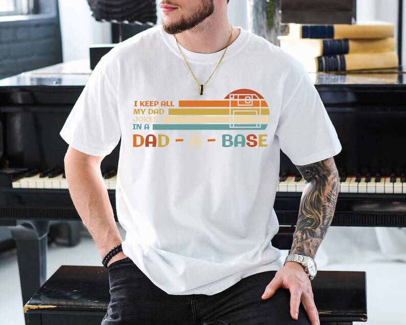 Puede incluir: Camiseta blanca con un gr&aacute;fico de estilo retro que dice "I keep all my dad jokes in a dad-a-base". El gr&aacute;fico presenta una cinta de casete y un esquema de color arco&iacute;ris.