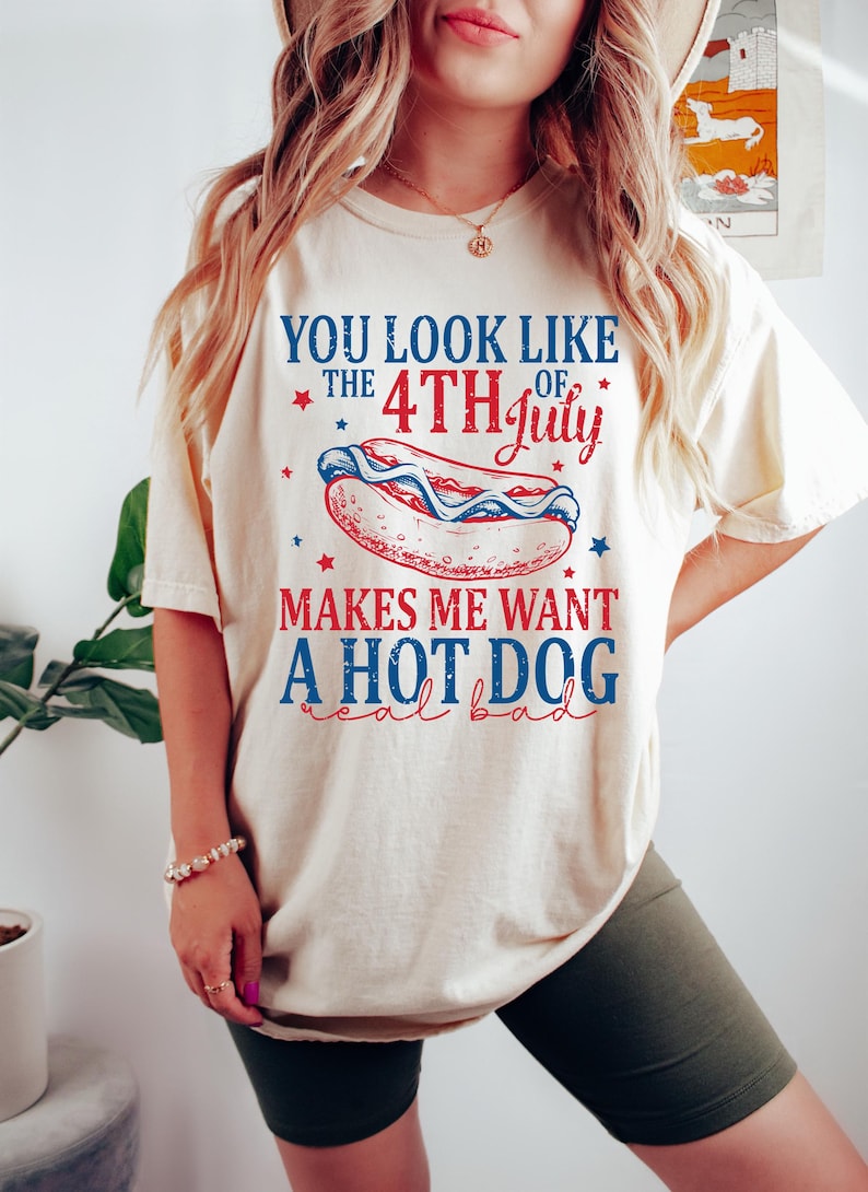 May include: Cream-colored t-shirt with a Fourth of July theme. The shirt features a graphic of a hot dog with red, white, and blue accents, and the text "You look like the 4th of July makes me want a hot dog real bad."
