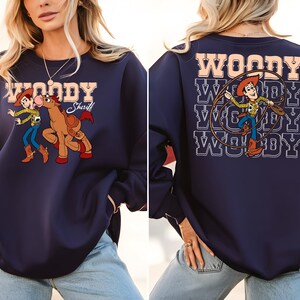 Puede incluir: Sudadera azul marino con un gr&aacute;fico de dibujos animados de Woody y un caballo en la parte delantera. La parte posterior de la sudadera tiene un gr&aacute;fico de Woody con un lazo y la palabra "WOODY" repetida. La sudadera tiene mangas largas.