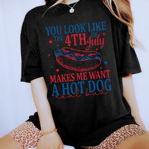 May include: Black t-shirt with a Fourth of July hot dog graphic. The text reads "You look like the 4th of July makes me want a hot dog real bad" in red and blue. The design includes stars and a hot dog illustration.