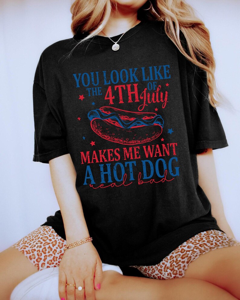 May include: Black t-shirt with a Fourth of July hot dog graphic. The text reads "You look like the 4th of July makes me want a hot dog real bad" in red and blue. The design includes stars and a hot dog illustration.