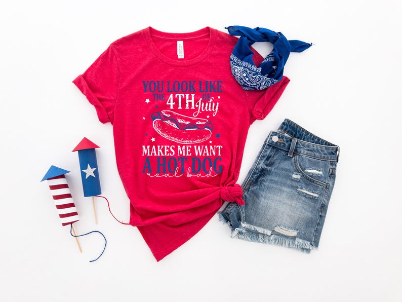 May include: A red t-shirt with the text "You look like the 4th of July makes me want a hot dog real bad" is paired with denim shorts and a blue bandana. Two decorative firework models complete the patriotic theme.