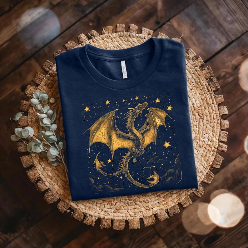 May include: A navy blue t-shirt featuring a golden dragon design with outstretched wings and stars. The dragon is intricately detailed, set against a starry night sky. The shirt is folded on a woven, circular placemat.