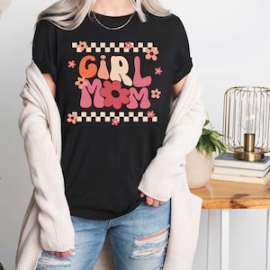 May include: A black t-shirt with a retro-style graphic that says "Girl Mom" in pink and red letters with flowers. The shirt is being worn by a person with a white cardigan.