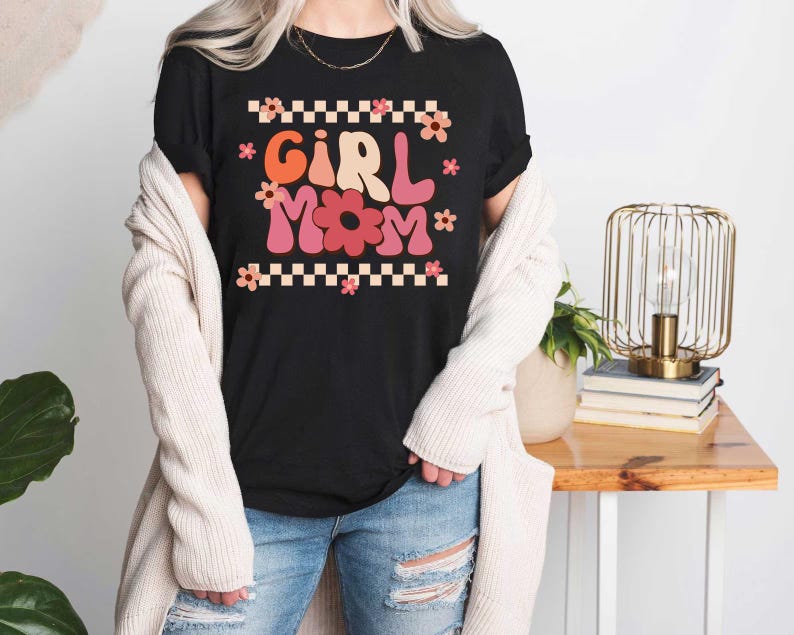 May include: A black t-shirt with a retro-style graphic that says "Girl Mom" in pink and red letters with flowers. The shirt is being worn by a person with a white cardigan.