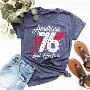 May include: Navy blue t-shirt with the text "America 76 land of the free" in white and red. The number 76 is filled with blue stars. The shirt is paired with denim shorts and brown sandals.
