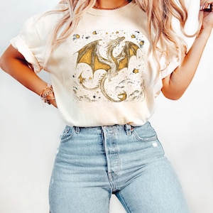 May include: Cream-colored t-shirt featuring a detailed illustration of a dragon with golden wings, set against a backdrop of clouds and stars. The dragon design is centered on the shirt, creating a fantasy-themed aesthetic. The shirt is paired with light blue denim jeans.