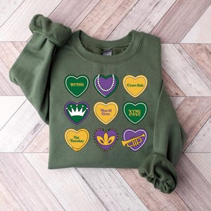 Peut inclure: Sweat-shirt vert sur le th&egrave;me du Mardi Gras. Le sweat-shirt comporte neuf c&oelig;urs avec diff&eacute;rents symboles et textes du Mardi Gras, notamment "beads", "crawfish", "Mardi Gras", "King Cake", "Fat Tuesday" et une couronne.