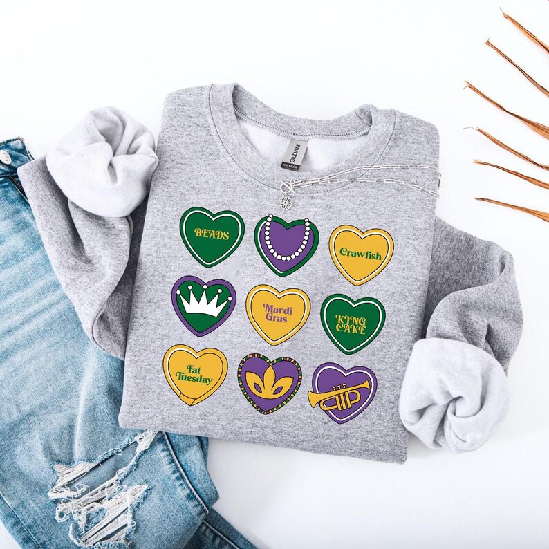 Peut inclure: Sweat-shirt gris avec un motif de neuf c&oelig;urs repr&eacute;sentant des textes et des images sur le th&egrave;me du Mardi Gras. Les c&oelig;urs sont verts, jaunes, violets et blancs. Le texte comprend "Bends", "Crawfish", "Mardi Gras", "King Cake", "Fat Tuesday", et "Laissez les bons temps rouler".