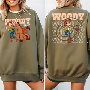 Puede incluir: Sudadera verde oliva con un gr&aacute;fico de dibujos animados de Woody y un caballo en la parte delantera. La parte posterior de la sudadera tiene un gr&aacute;fico de Woody con un lazo y la palabra "WOODY". La sudadera tiene cuello redondo y mangas largas.