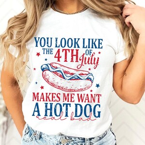 May include: White t-shirt with a patriotic design. The shirt features text that reads "YOU LOOK LIKE THE 4TH OF JULY MAKES ME WANT A HOT DOG real bad" with a hot dog graphic in red, white, and blue.