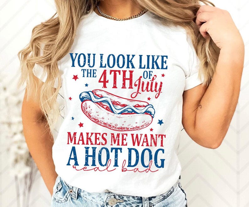 May include: White t-shirt with a patriotic design. The shirt features text that reads "YOU LOOK LIKE THE 4TH OF JULY MAKES ME WANT A HOT DOG real bad" with a hot dog graphic in red, white, and blue.