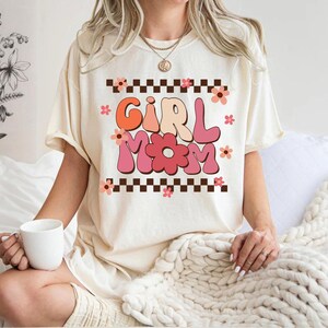 May include: A white t-shirt with a pink and orange graphic that says "Girl Mom" with flowers and a checkered pattern.