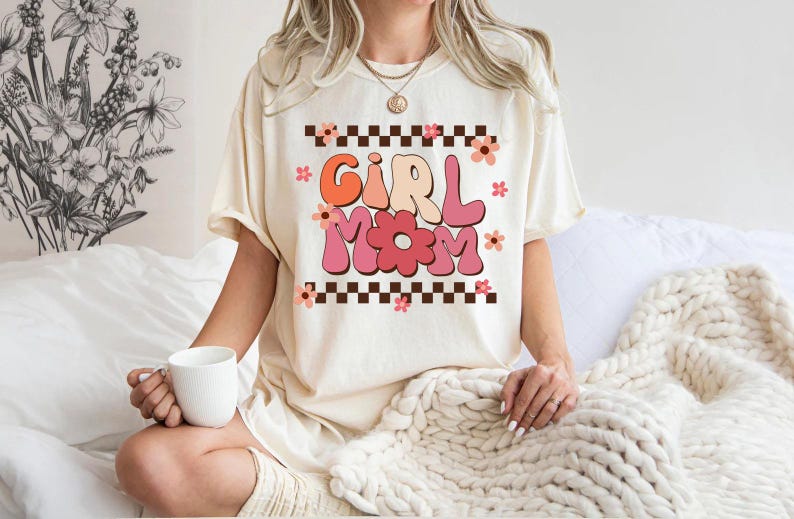 May include: A white t-shirt with a pink and orange graphic that says "Girl Mom" with flowers and a checkered pattern.