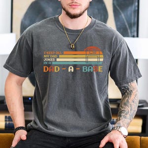 Puede incluir: Una camiseta gris oscura con un gr&aacute;fico de estilo vintage que dice "I keep all my dad jokes in a dad-a-base". El gr&aacute;fico presenta una cinta de casete de estilo retro y rayas de colores.