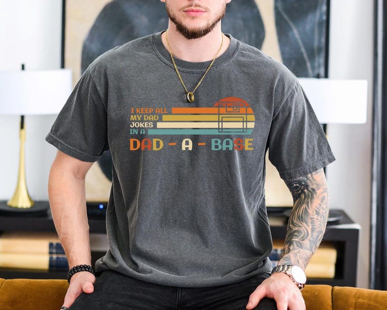 Puede incluir: Una camiseta gris oscura con un gr&aacute;fico de estilo vintage que dice "I keep all my dad jokes in a dad-a-base". El gr&aacute;fico presenta una cinta de casete de estilo retro y rayas de colores.