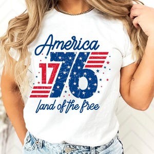 May include: White t-shirt with a patriotic design. The shirt features the word "America" in blue script above the number "76" in blue with white stars. Red and white stripes and the phrase "land of the free" complete the design.