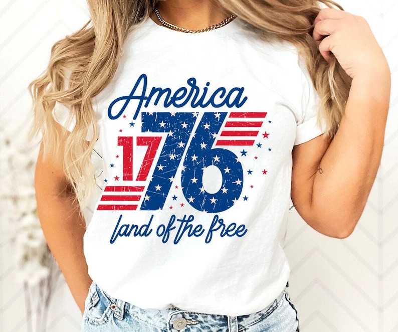 May include: White t-shirt with a patriotic design. The shirt features the word "America" in blue script above the number "76" in blue with white stars. Red and white stripes and the phrase "land of the free" complete the design.
