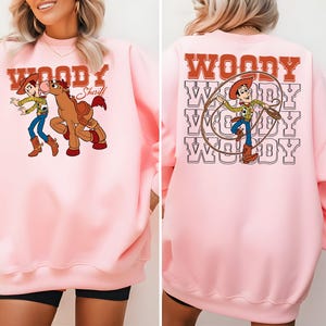 Puede incluir: Sudadera rosa claro con un dise&ntilde;o de dibujos animados de Woody y un caballo. La parte delantera de la sudadera tiene la palabra "Woody" encima del dise&ntilde;o, y la parte trasera tiene la palabra "Woody" repetida varias veces con un dise&ntilde;o de Woody.