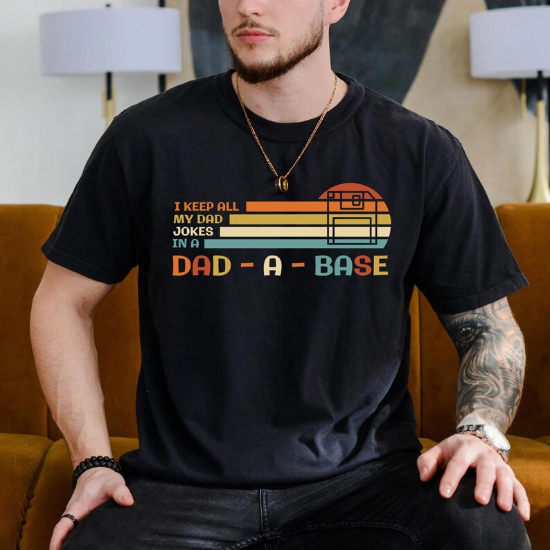 Puede incluir: Una camiseta negra con un dise&ntilde;o retro que presenta un disquete y el texto "I keep all my dad jokes in a dad-a-base".
