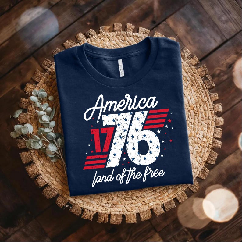 May include: A navy blue t-shirt featuring the text "America 1776 land of the free" in white and red. The design includes the number 76 with white stars and red stripes. The shirt is folded on a woven mat, evoking patriotic themes.