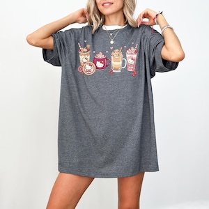 May include: Dark grey t-shirt with a colourful graphic of various drinks, including coffee and milkshakes, with a cartoon character design. The design includes hearts and berries. The shirt has short sleeves and a crew neck.