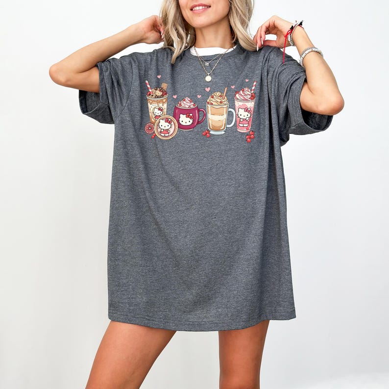 May include: Dark grey t-shirt with a colourful graphic of various drinks, including coffee and milkshakes, with a cartoon character design. The design includes hearts and berries. The shirt has short sleeves and a crew neck.