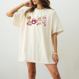 May include: Cream-coloured oversized t-shirt featuring a colourful graphic of various beverages, including coffee and milkshakes, with a cartoon character design. The design includes hearts and cherries. The shirt has short sleeves and a relaxed fit.