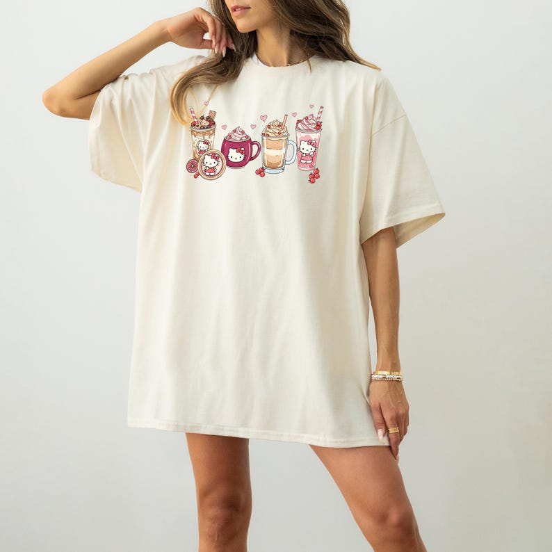 May include: Cream-coloured oversized t-shirt featuring a colourful graphic of various beverages, including coffee and milkshakes, with a cartoon character design. The design includes hearts and cherries. The shirt has short sleeves and a relaxed fit.