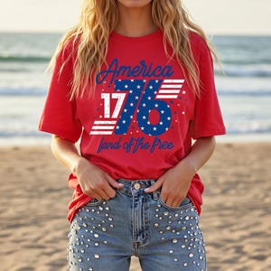 May include: A red t-shirt with the text "America 1776 land of the free" in blue and white. The design includes stars and stripes. The model is wearing blue jeans with decorative embellishments.