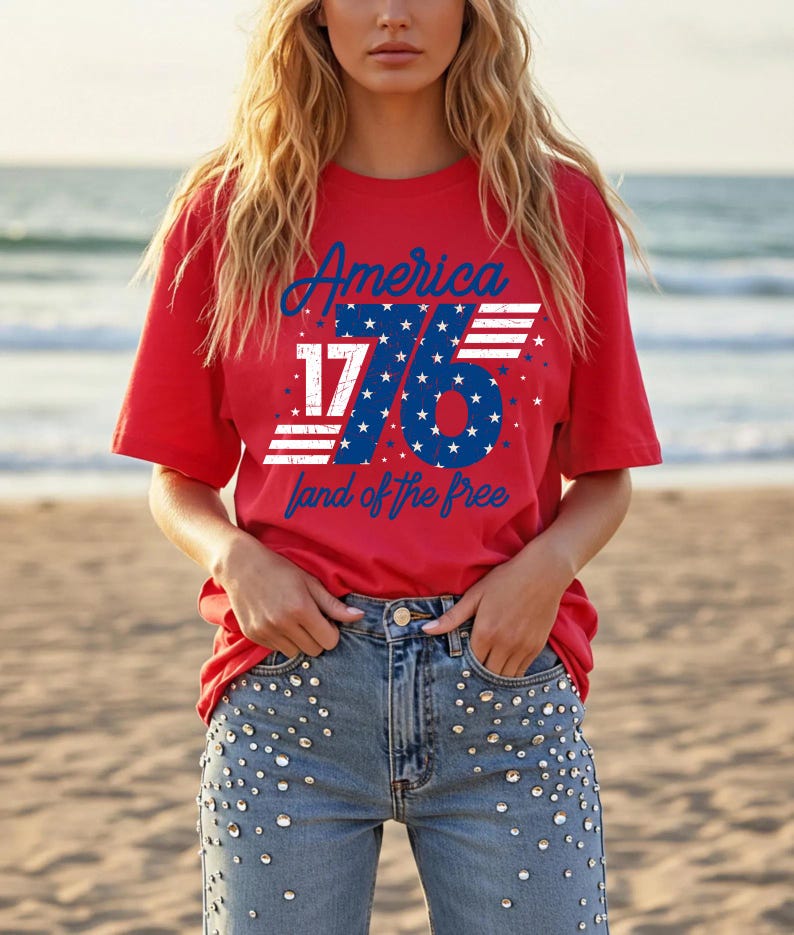 May include: A red t-shirt with the text "America 1776 land of the free" in blue and white. The design includes stars and stripes. The model is wearing blue jeans with decorative embellishments.