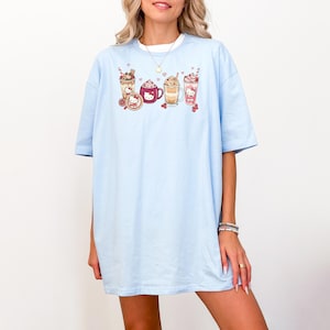 May include: Pale blue t-shirt with a graphic of cartoon beverages, such as coffee and milkshakes. The design includes a cartoon character. The shirt has short sleeves and a loose fit, ideal for everyday wear.