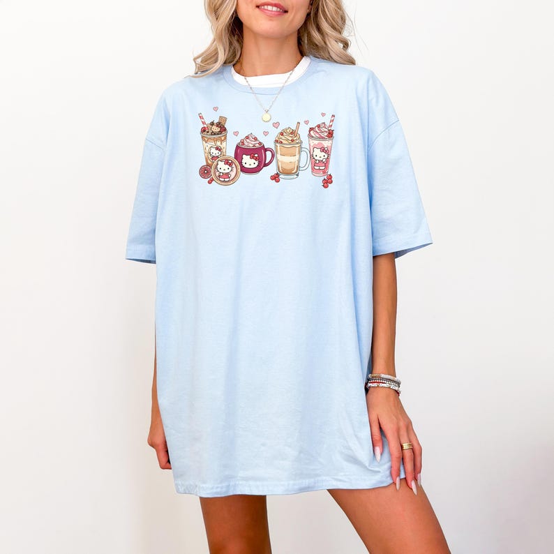May include: Pale blue t-shirt with a graphic of cartoon beverages, such as coffee and milkshakes. The design includes a cartoon character. The shirt has short sleeves and a loose fit, ideal for everyday wear.