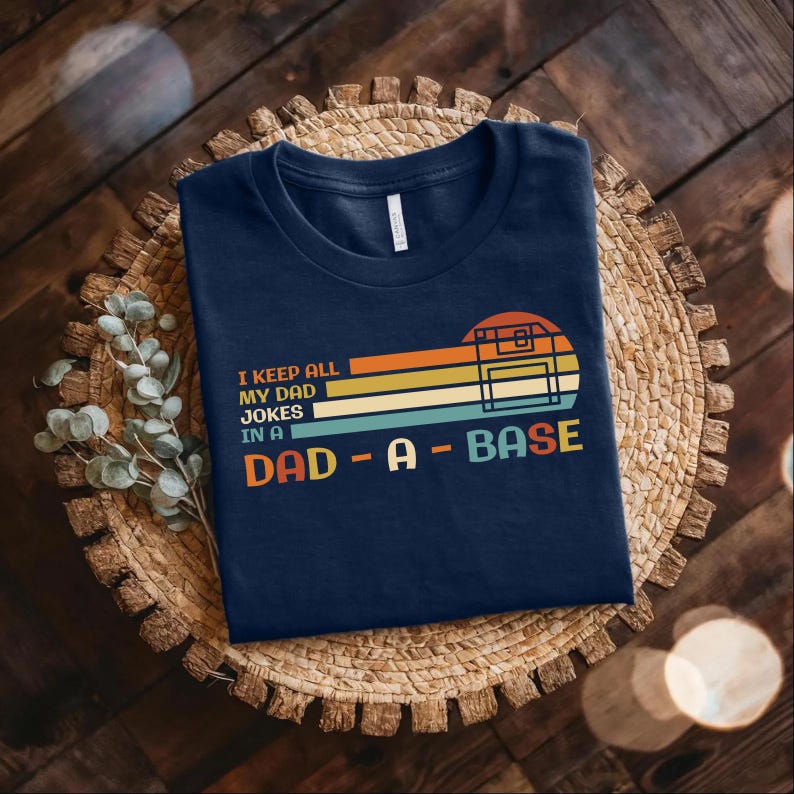 Puede incluir: Una camiseta azul marino con un gr&aacute;fico retro de un disquete y el texto "I keep all my dad jokes in a dad-a-base".