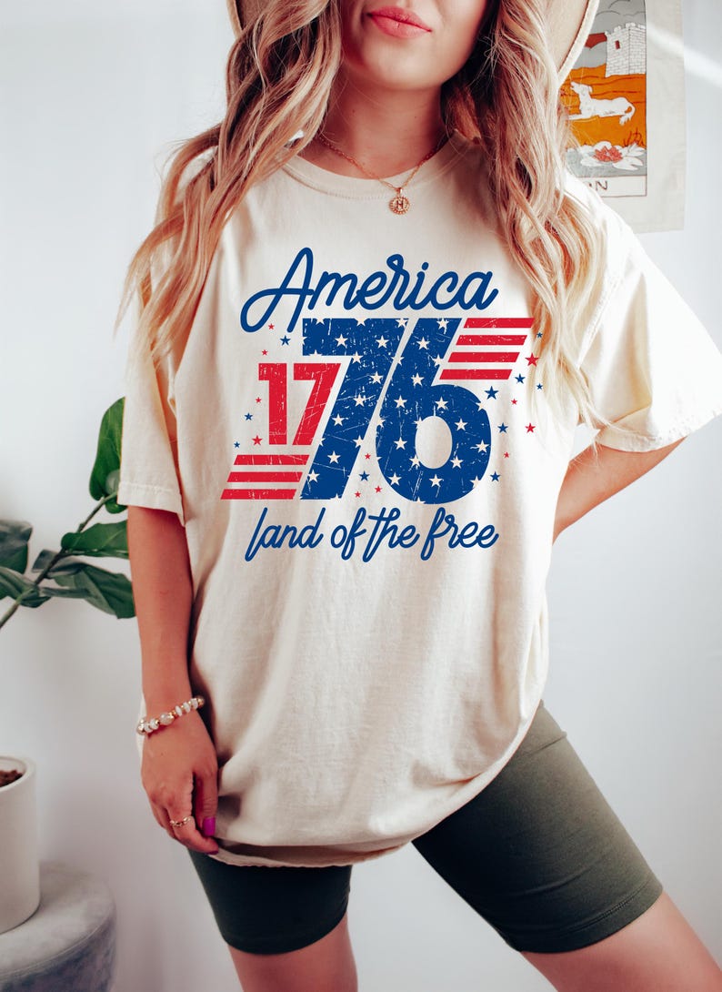 May include: A cream-coloured t-shirt with a patriotic design. The design features the word "America" in blue script above the number "1776" in red and blue, with the phrase "land of the free" below. The shirt is a casual style.