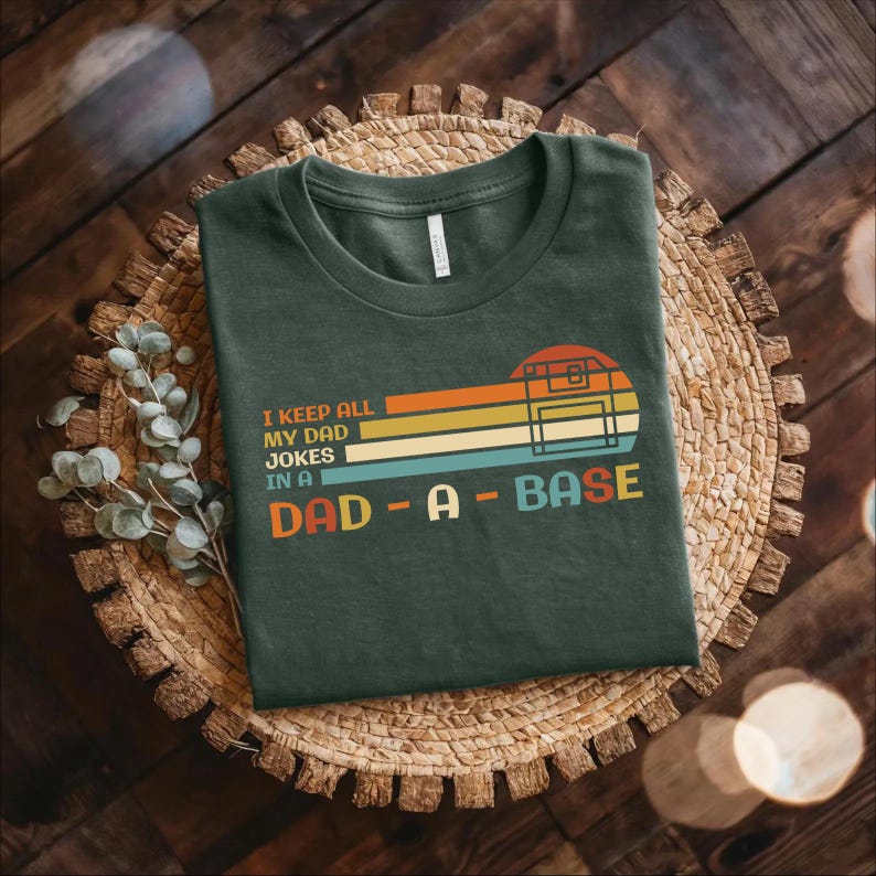 Puede incluir: Una camiseta verde oscuro con un gr&aacute;fico de estilo retro que dice "I keep all my dad jokes in a dad-a-base". El gr&aacute;fico presenta un disquete con un fondo de arco&iacute;ris.