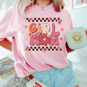 May include: Pink t-shirt with a retro-style graphic that says "Girl Mom" in colorful letters with flowers and a checkered border.