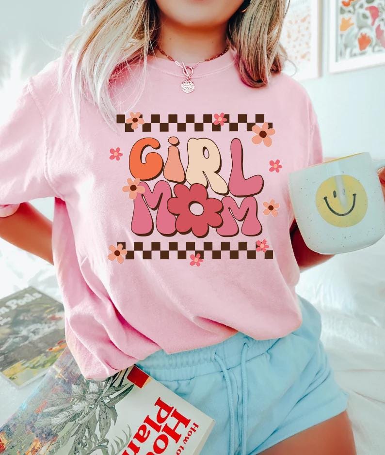 May include: Pink t-shirt with a retro-style graphic that says "Girl Mom" in colorful letters with flowers and a checkered border.