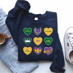Peut inclure: Un sweat-shirt bleu marine sur le th&egrave;me du Mardi Gras. Le sweat-shirt pr&eacute;sente une vari&eacute;t&eacute; de motifs en forme de c&oelig;ur avec des textes tels que "Beads", "Mardi Gras", "King Cake", "Fat Tuesday" et "Crawfish".