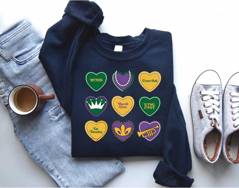 Peut inclure: Un sweat-shirt bleu marine sur le th&egrave;me du Mardi Gras. Le sweat-shirt pr&eacute;sente une vari&eacute;t&eacute; de motifs en forme de c&oelig;ur avec des textes tels que "Beads", "Mardi Gras", "King Cake", "Fat Tuesday" et "Crawfish".