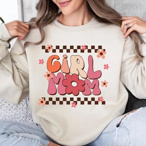 May include: A cream-colored sweatshirt with a graphic design of the words "Girl Mom" in a retro font with pink, orange, and brown colors. The graphic is surrounded by a checkered pattern and pink flowers.