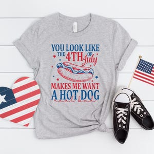May include: A gray t-shirt with the text "YOU LOOK LIKE THE 4TH OF JULY MAKES ME WANT A HOT DOG real bad." The image also includes a heart-shaped decoration, a small American flag, and black sneakers.