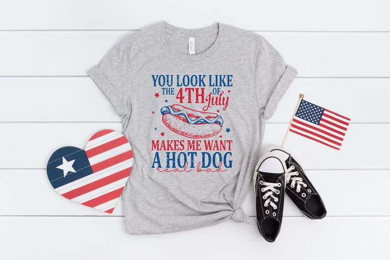 May include: A gray t-shirt with the text "YOU LOOK LIKE THE 4TH OF JULY MAKES ME WANT A HOT DOG real bad." The image also includes a heart-shaped decoration, a small American flag, and black sneakers.