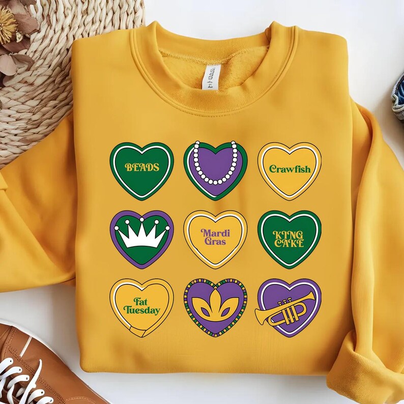Peut inclure: Un sweat-shirt jaune avec un motif de neuf c&oelig;urs, chacun avec un th&egrave;me diff&eacute;rent du Mardi Gras. Les c&oelig;urs pr&eacute;sentent des textes comme "Beads", "Crawfish", "King Cake", "Mardi Gras" et "Fat Tuesday".