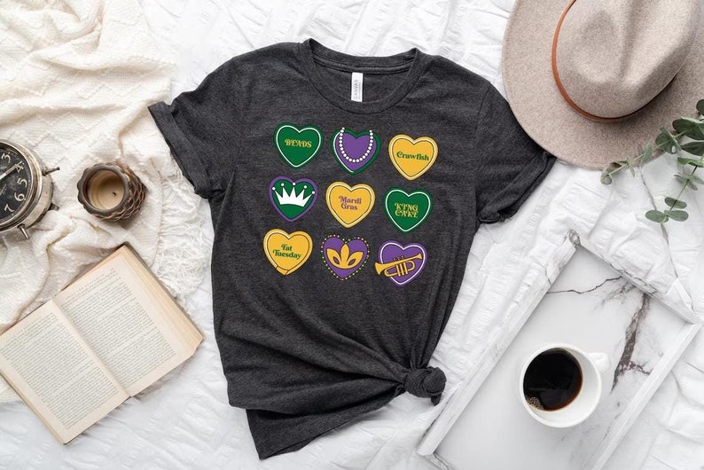 Peut inclure: Un t-shirt gris fonc&eacute; avec un motif de neuf c&oelig;urs violets, verts et jaunes. Chaque c&oelig;ur a un design et un texte diff&eacute;rents, notamment "Bends", "King Cake", "Mardi Gras" et "Fat Tuesday".