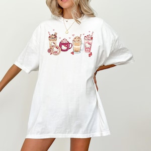 May include: White t-shirt with a graphic of various drinks, such as a milkshake, coffee, and hot chocolate, decorated with cute character designs. The design includes hearts and cherries. The shirt has a relaxed, oversized fit.
