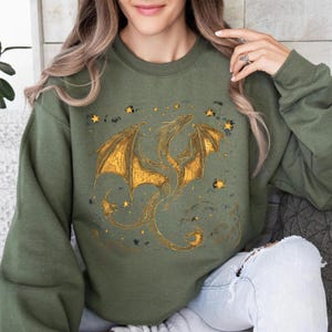 May include: Olive green sweatshirt featuring a golden dragon design with outstretched wings and star accents. The dragon is intricately detailed, with a textured appearance. The design is centered on the front of the sweatshirt, creating a fantasy-themed aesthetic.